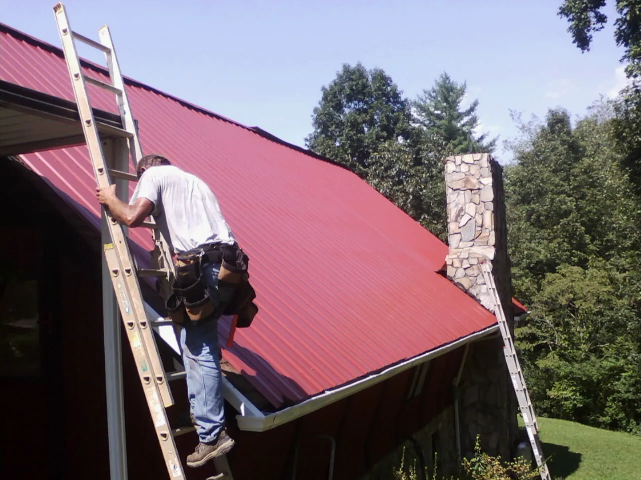 Roofer on a ladder beside a red metal roof and stone chimney — Soffit & Fascia Repair by Roofing Brothers in Martins Ferry